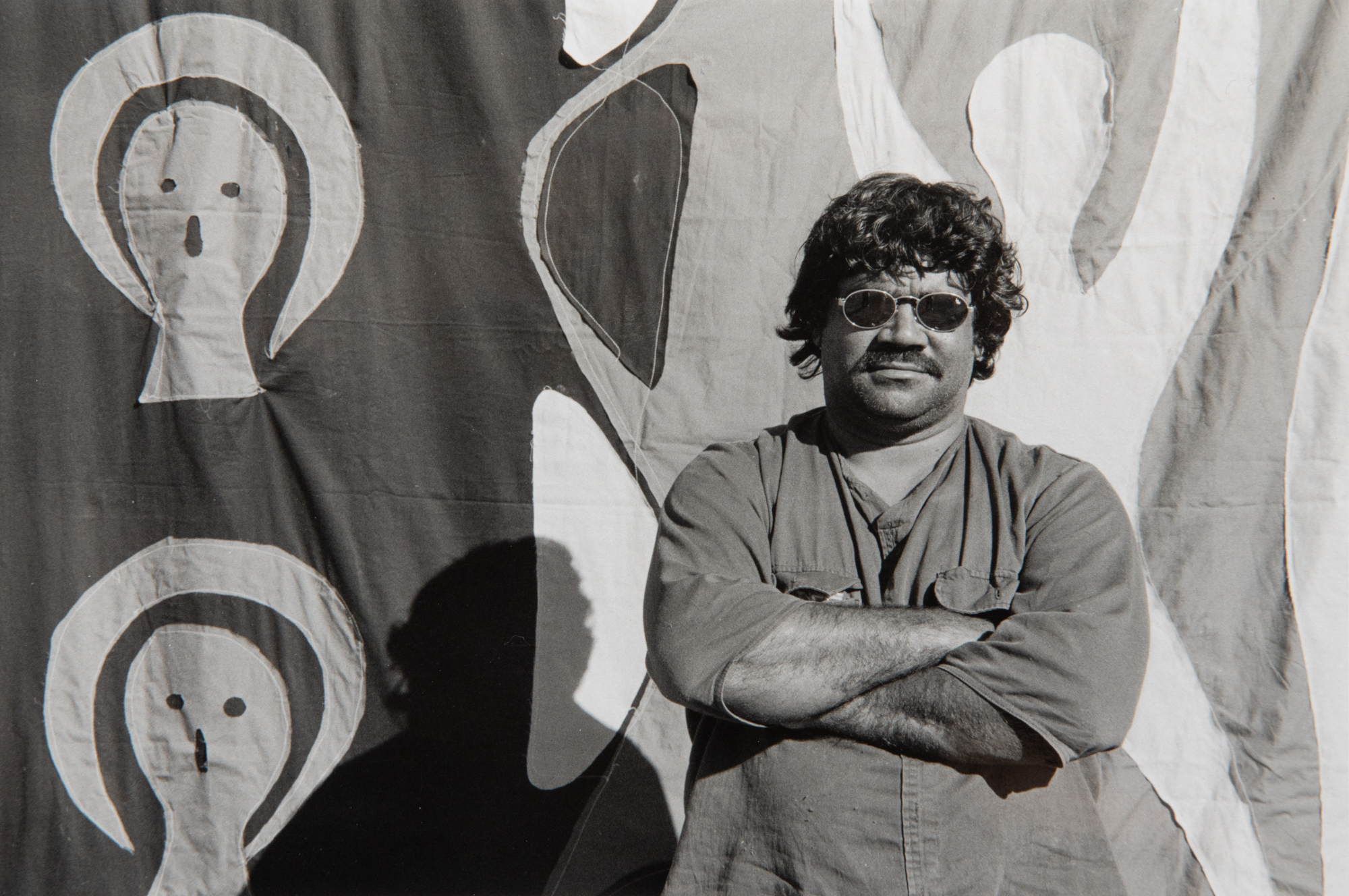 Black and white photo of Anthony standing with arms crossed, wearing sunglasses in front of a cloth stitched mural 