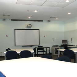 There are desks set up in a square in f the room. The front of the room has a whiteboard and a projector set up.