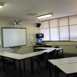 There are desks set up in a u-shape facing the front of the room. The front of the room has a whiteboard and a projector set up.