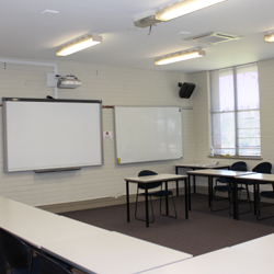 There are desks set up in a u-shape facing the front of the room. The front of the room has a whiteboard and a projector set up.