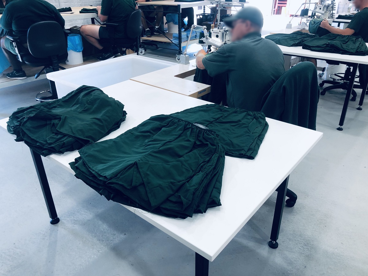 Table with 3 piles of green shorts in foreground, and the back of a man in a green shirt sitting ata sewing machine