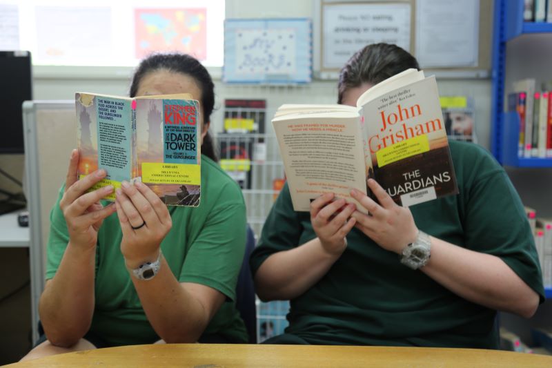 Two seated women, dressed in green t-shirts, holding books up in front of their faces  