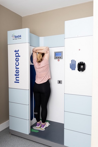 A woman stands in the scanner facing sideways with her legs together, arms raised above her head.
